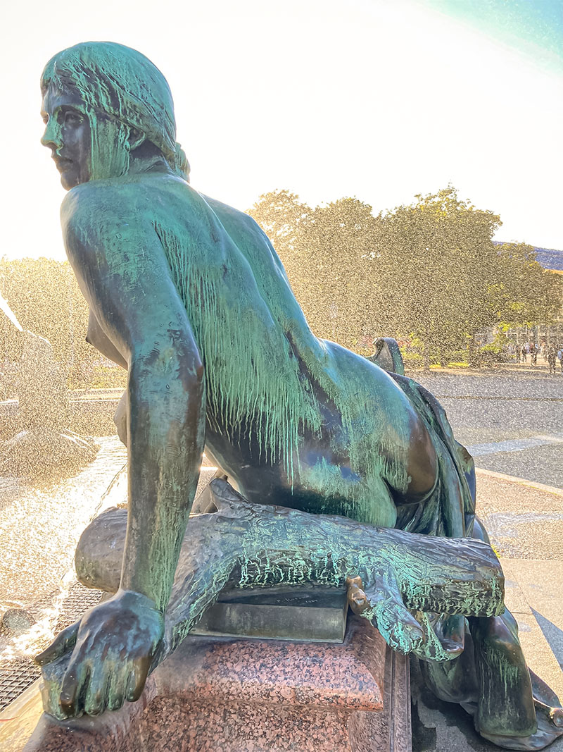 Bronzestatue sitzend auf einem Arm abgestützt an einem Brunnen, Wassertropfen in der Luft, sommerliche Stimmung